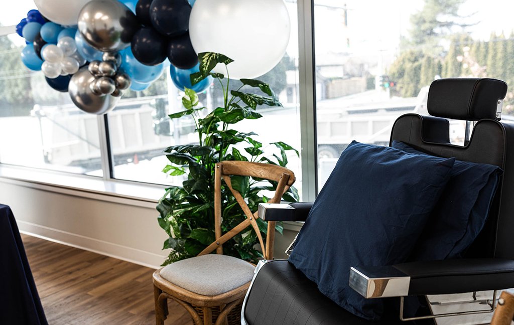 Black leather chair with navy pillows beside a wooden chair, a plant and blue and silver balloons.