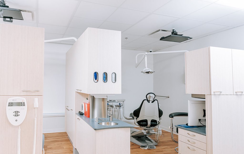 Dental treatment area with a patient chair, overhead light, light wood cabinets and a sink.