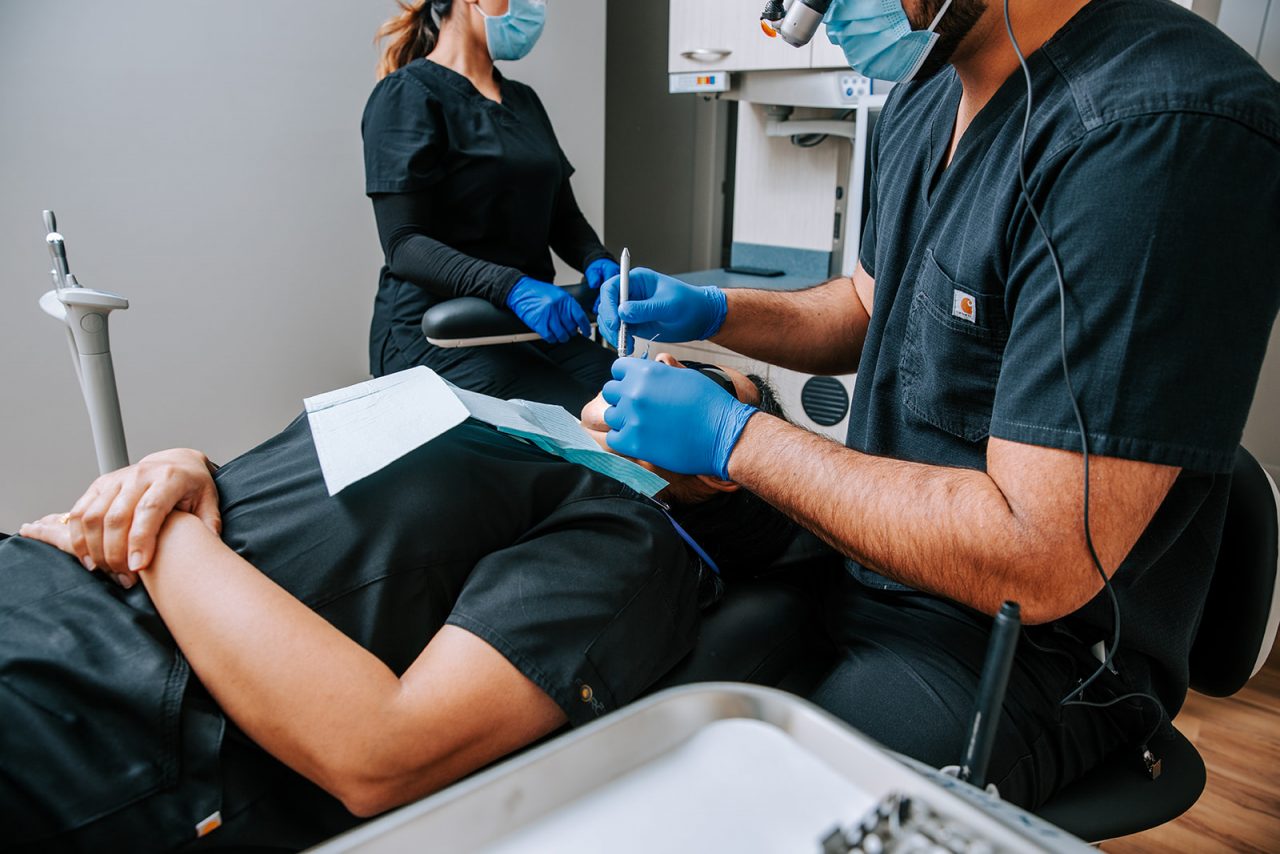 Dentist in dark scrubs and blue gloves working on a patient lying in a dental chair.