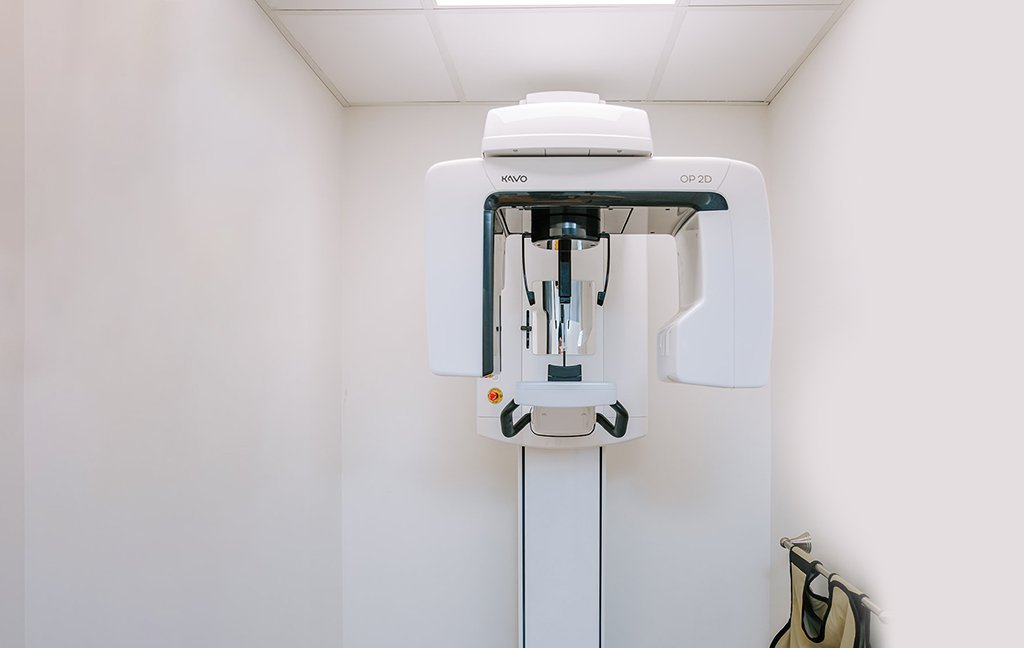 Panoramic dental X-ray machine in a small white room with a lead apron on a hook.