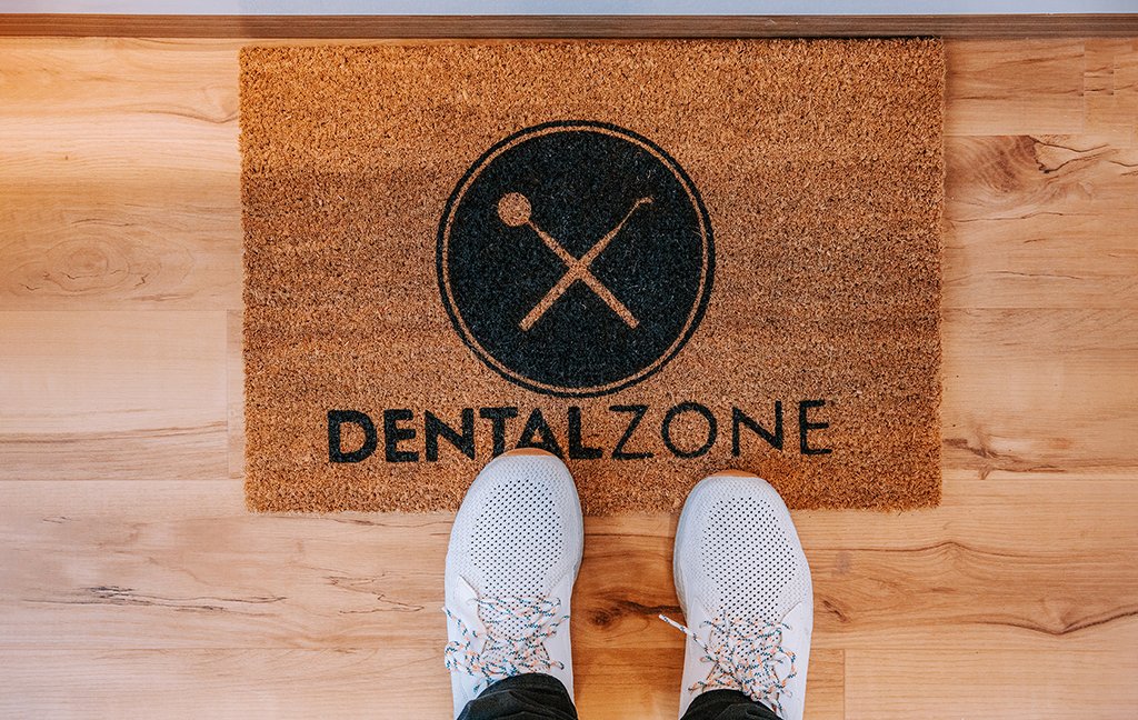 Top-down view of a Dental Zone logo doormat on a wood floor, with a person's white sneakers.