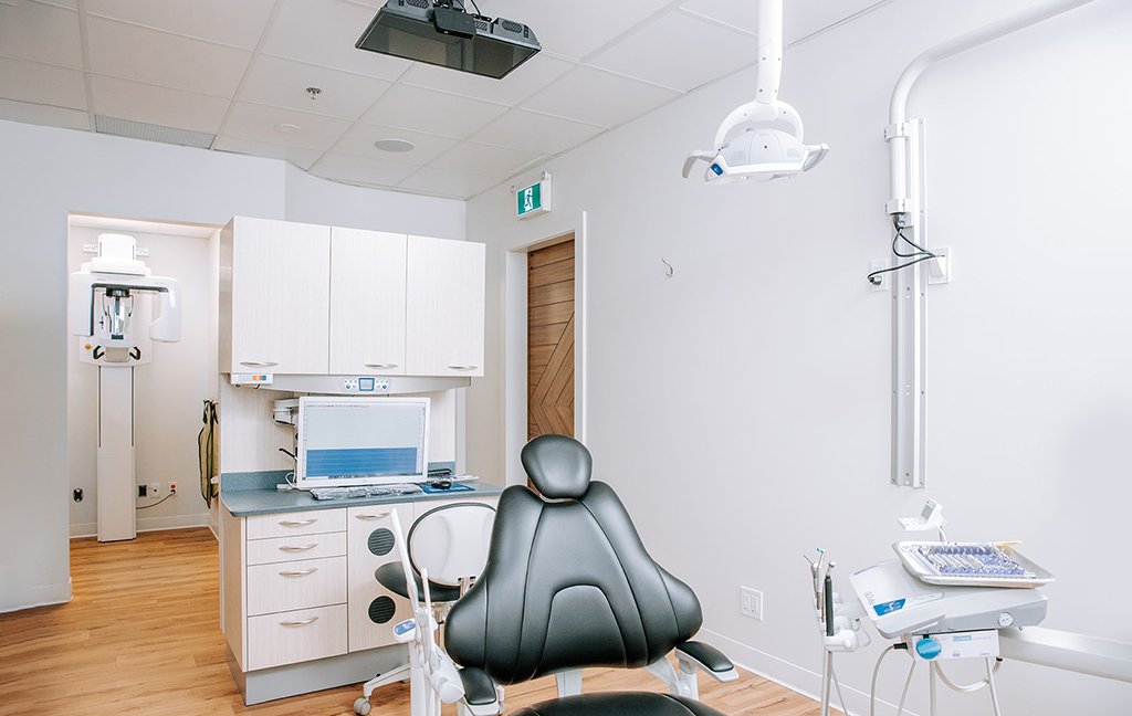 Treatment room with a black dental chair, overhead light, cabinets and a monitor, and an X-ray machine beyond.