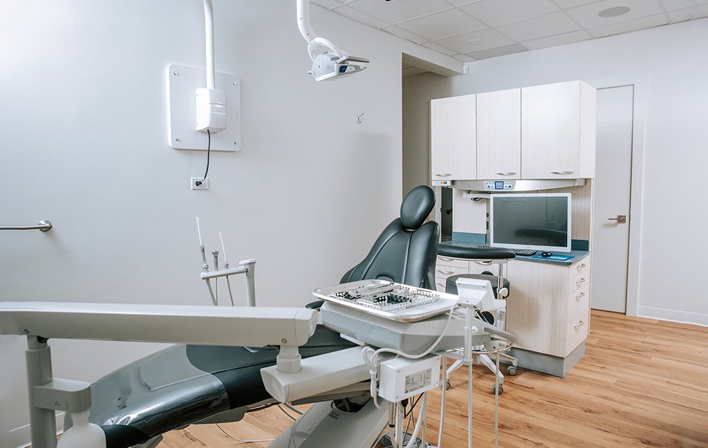 Treatment room with a dental chair, instrument tray, wall light, cabinets and a computer monitor.