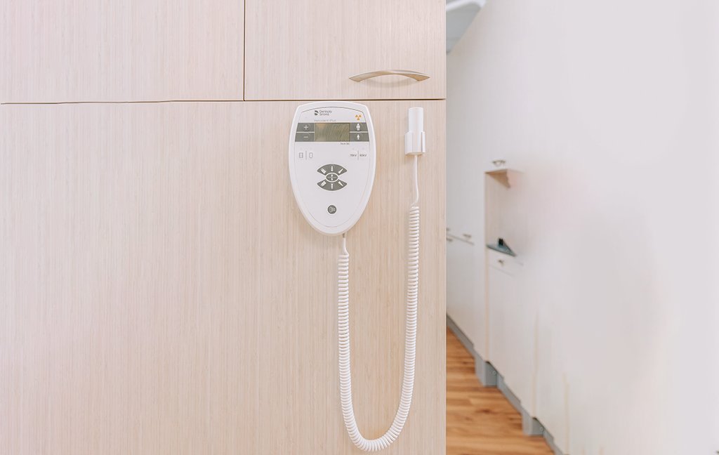 Wall-mounted dental X-ray control panel with a coiled cord on a light wood cabinet.